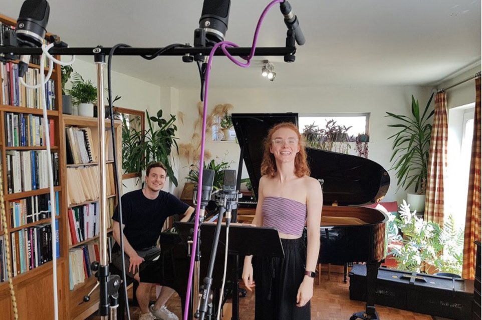 Two musicians, a pianist and a singer in a recording studio; there is a piano, sheet music stand, microphones and other recording gears.